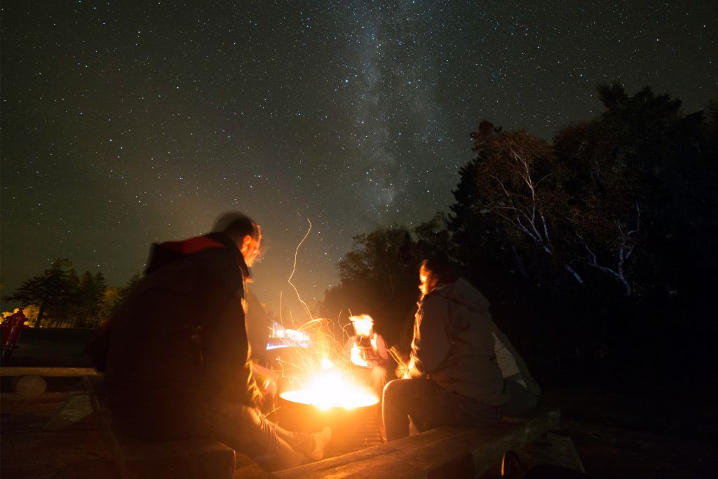 group of three around a bond fire in a Maine starry night backdrop