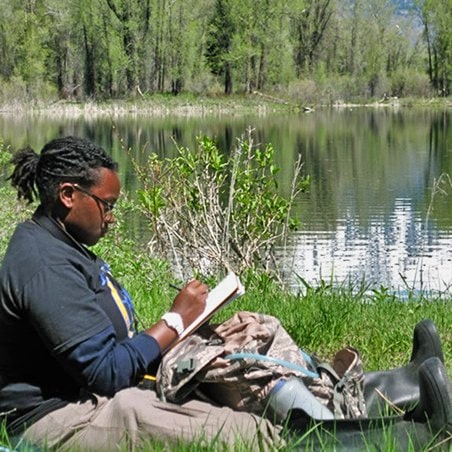 A young man sketching alongside a lake and trees.