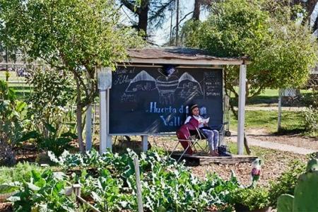 A girl sitting in front of the Huerta del Valle community garden banner.